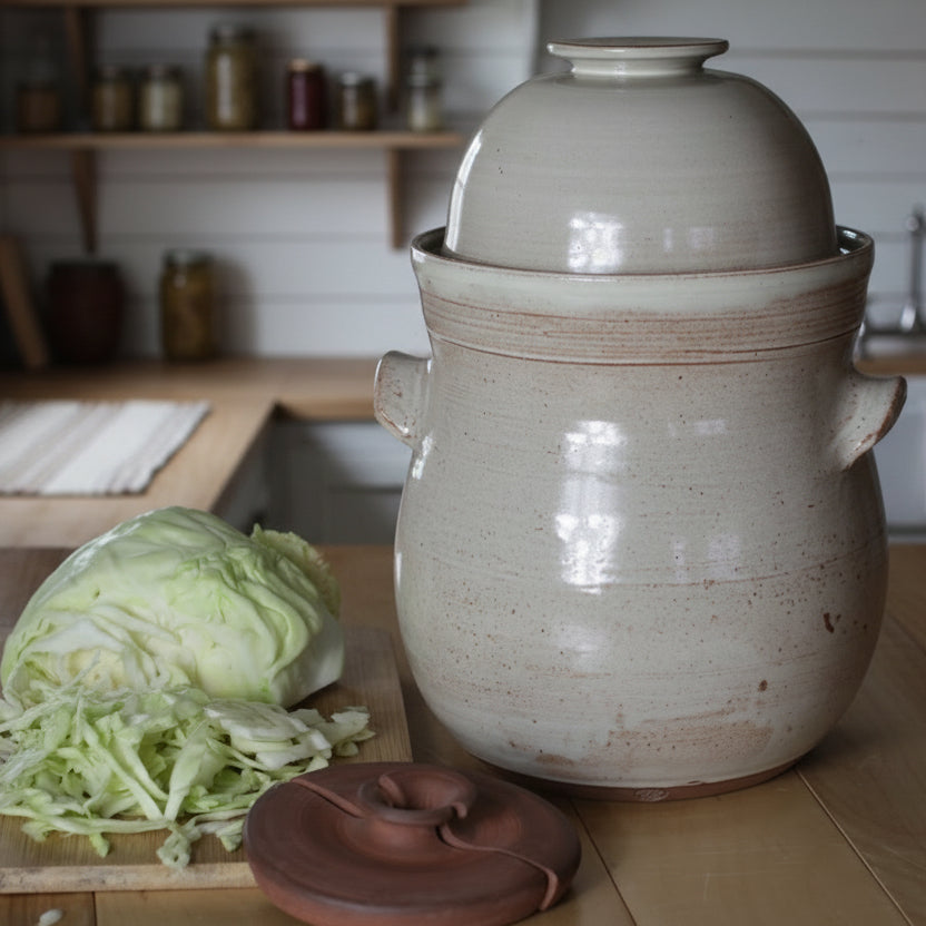 White ceramic crock with a lid on a wooden surface next to a head of cabbage and a cutting board.